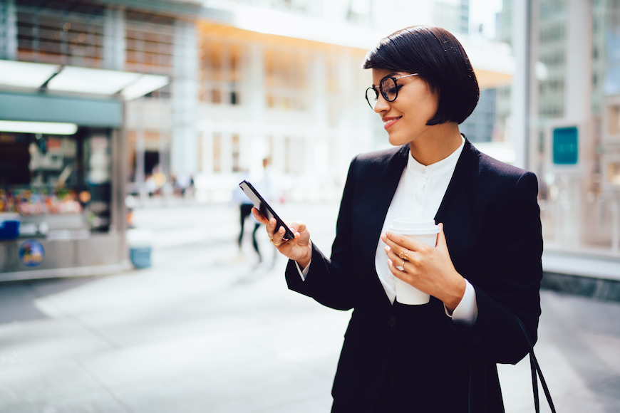 Female business-person checking their phone while holding a travel coffee cup.