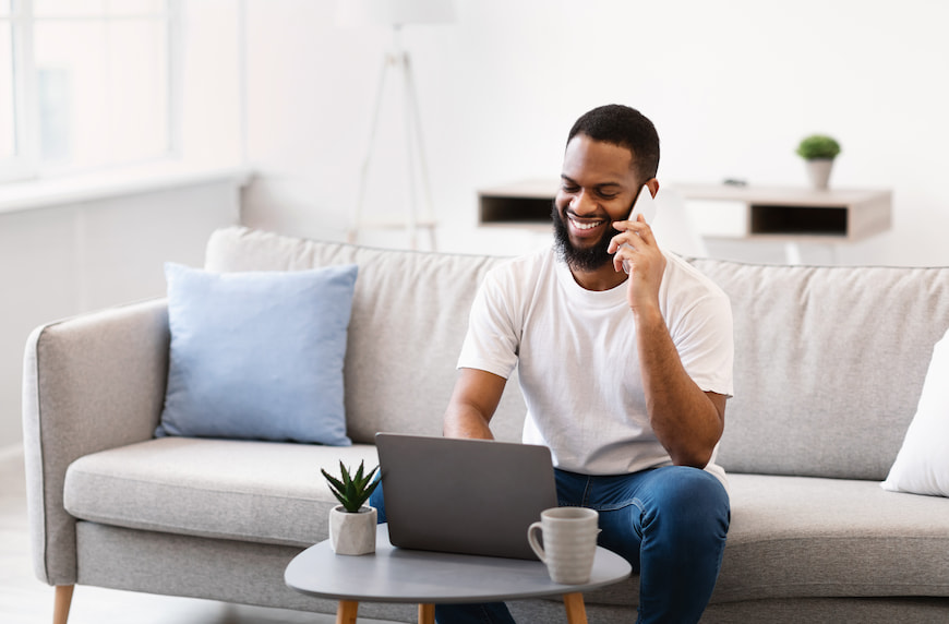 Man sitting on couch using the internet on his computer while talking on his cell phone